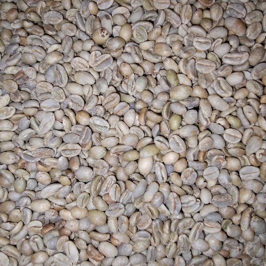 A close-up view of many raw coffee beans filling the frame, showing their light tan to beige color with subtle greenish and brown variations.