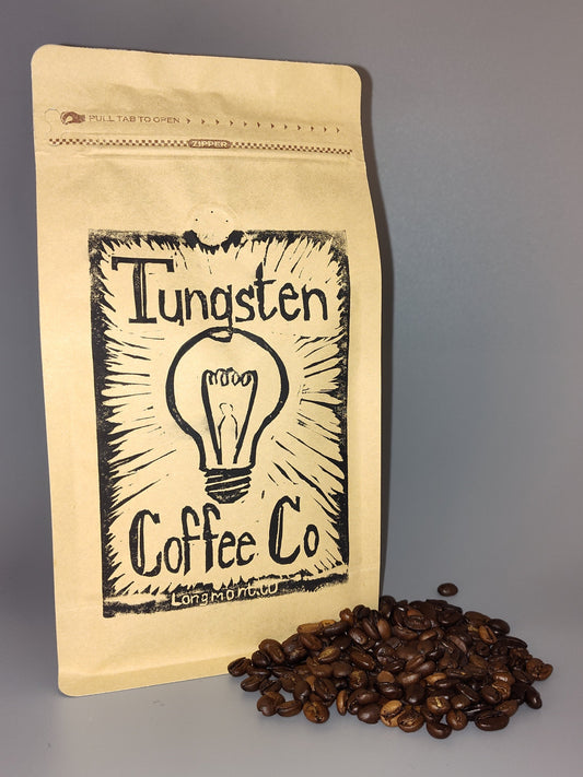 A kraft paper stand-up pouch labeled 'Tungsten Coffee Co' with a black sketch-style lightbulb logo, standing next to a small pile of dark roasted coffee beans on a smooth gray surface.